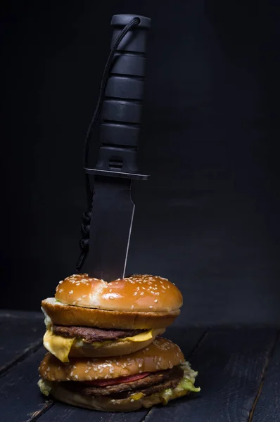 Μαύρο μεγάλο μαχαίρι εισάγεται ένα burger. Μαύρο φόντο. — Φωτογραφία Αρχείου