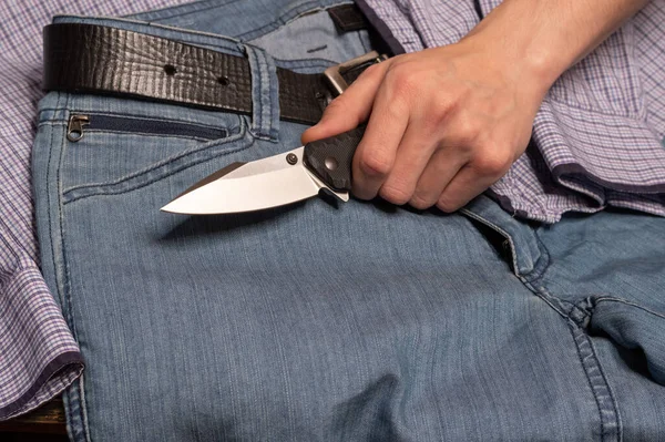 El cuchillo en la mano sobre el fondo de los vaqueros y la camisa. Ropa casual para hombre y un cuchillo plegable en su mano . — Foto de Stock