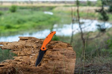 Turuncu saplı ve siyah bıçaklı bir bıçak. Nehrin arka planında eski bir ağaç kütüğünde bıçak..