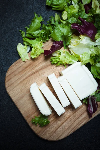 Greek feta cheese on a wooden board, lettuce leaves on a black b — Stock Photo, Image