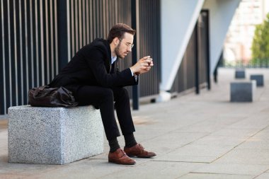 Takım elbiseli genç işadamı oturup telefonda oynuyor. Sosyal ağlar bağımlılığı. Mobil uygulama kullanılıyor.