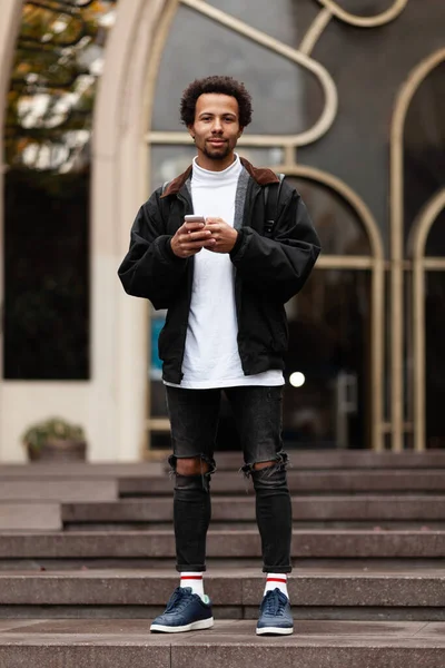 Attractive young african male hipster in ripped jeans with phone in his hands on  street.