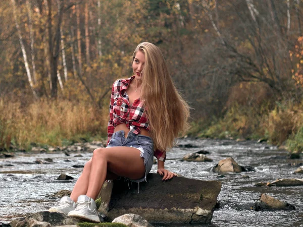 穿着短裤和格子衬衫的苗条运动金发女郎坐在河中央的石头上. — 图库照片