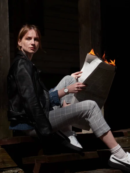 Deri ceketli, damalı pantolonlu ve elinde yanan bir gazete olan beyaz spor ayakkabılı genç bir kadın..