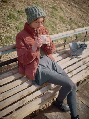 Gri etek ve külotlu çorap giyen, yeşil bir şapka ve kırmızı ceket giyen bir kadın, bankta oturur ve çay içer..