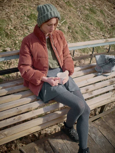 一位身着灰色裙子和裤袜、戴着绿色帽子、穿着红色夹克的妇女坐在长凳上喝茶. — 图库照片
