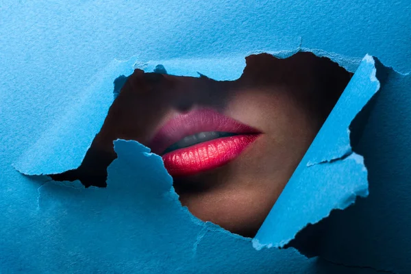 Uma visão dos lábios femininos através de um buraco em um fundo azul papel — Fotografia de Stock