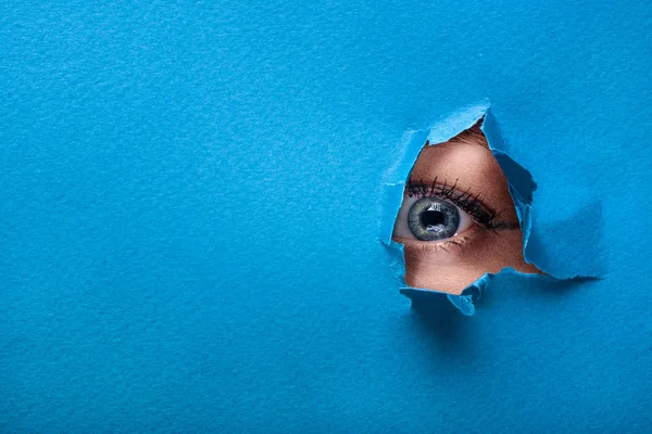 Um olho feminino olha através de um buraco em um papel fundo azul — Fotografia de Stock