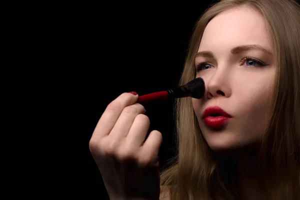 Portrait of a beautiful blonde who uses a makeup brush on a black background.