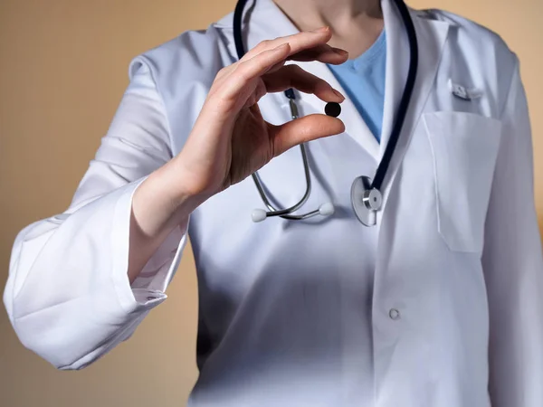 A női orvos a sztetoszkóppal egy bézs háttér tart egy fekete kerek tabletta. — Stock Fotó