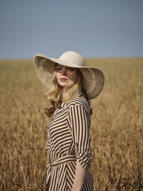 Buğday tarlasında ki bir sarışın hasır şapka ve dikey çizgili bir elbiseyle yürüyor..