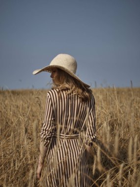 Buğday tarlasında ki bir sarışın hasır şapka ve dikey çizgili bir elbiseyle yürüyor..