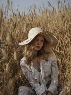 Beyaz bluz ve hasır şapkalı bir sarışın çavdar tarlasında oturur.