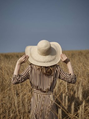 Buğday tarlasında ki bir sarışın hasır şapka ve dikey çizgili bir elbiseyle yürüyor..