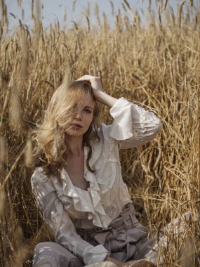 Beyaz bluz giymiş bir sarışın çavdar tarlasında oturur.