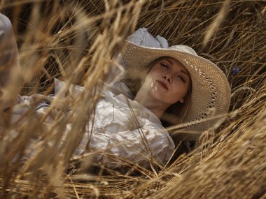 Beyaz bluz ve hasır şapkalı bir sarışın çavdar tarlasında yatıyor.