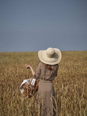 Bir buğday tarlasında bir sarışın kireç şapka ve dikey çizgili elbise elma bir hasır sepet ile yürüyor.