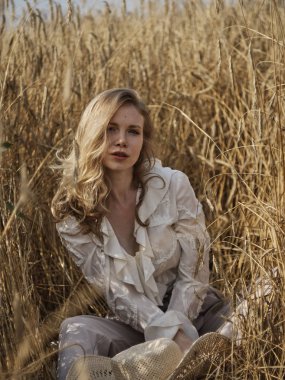 Beyaz bluz giymiş bir sarışın çavdar tarlasında oturur.