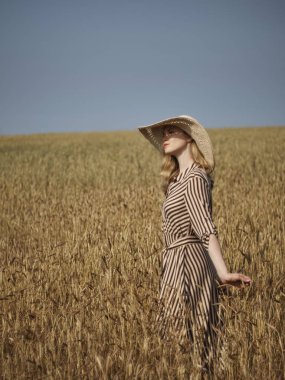 Buğday tarlasında ki bir sarışın hasır şapka ve dikey çizgili bir elbiseyle yürüyor..