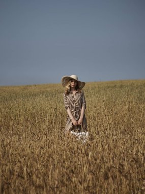 Bir buğday tarlasında bir sarışın kireç şapka ve dikey çizgili elbise elma bir hasır sepet ile yürüyor.