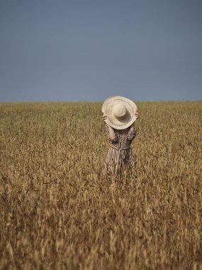 Buğday tarlasında ki bir sarışın hasır şapka ve dikey çizgili bir elbiseyle yürüyor..