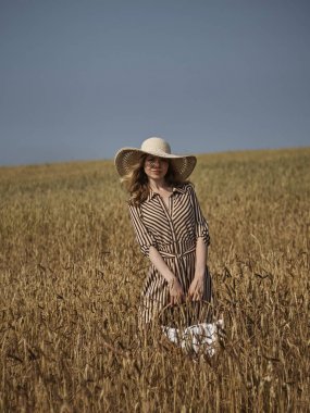Bir buğday tarlasında bir sarışın kireç şapka ve dikey çizgili elbise elma bir hasır sepet ile yürüyor.