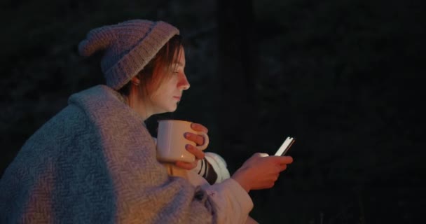 Una joven al atardecer se sienta en un bosque frente a un fuego y usa un teléfono inteligente . — Vídeo de stock