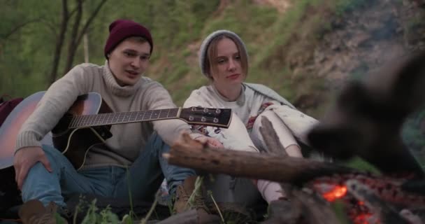 Um amigo com uma guitarra e namorada estão sentados na frente de uma fogueira na natureza e conversando diversão . — Vídeo de Stock