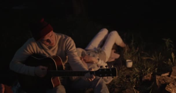 A young couple sits at night by the fire. The guy plays the guitar and his girlfriend lies on his lap and listens to music. — Stock Video