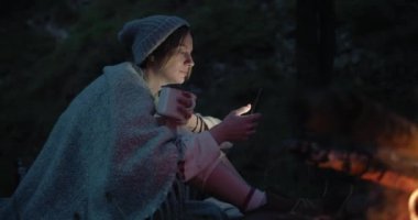 alacakaranlıkta genç bir kadın bir yangın karşısında bir ormanda oturur ve bir akıllı telefon kullanır. 