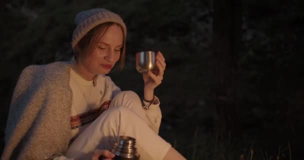 A young woman sits at night in a forest opposite a fire and pours tea from a thermos and drinks it. — Stock Video