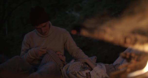 Um jovem casal senta-se à noite junto à lareira. O cara toca guitarra e sua namorada se deita em seu colo e ouve música . — Vídeo de Stock