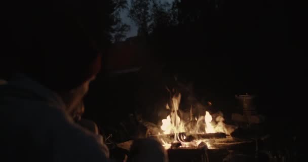 Egy fiatal pár ül éjjel az erdőben, magába foglaló közös kockás előtt egy tábortűz, beszéd és nevetve. — Stock videók