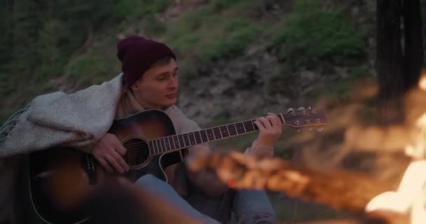 Ένας νεαρός κάθεται μπροστά σε μια φωτιά στην όχθη του ποταμού και παίζει την κιθάρα. — Αρχείο Βίντεο