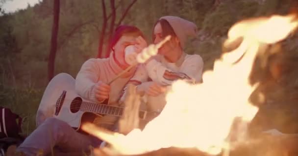 Ein junges Paar sitzt am Feuer und redet, ein Mann frittiert Marshmallows und trinkt Tee aus demselben Becher. — Stockvideo