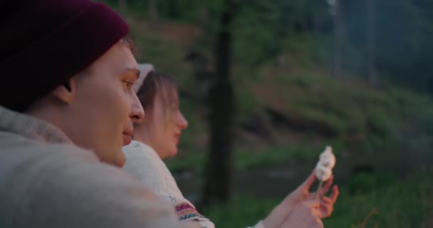 Genç bir çift bir dağ nehrinin kıyısında bir ormanda oturur, sohbet ve marshmallow kızartma. — Stok video