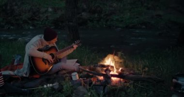genç bir adam nehir kıyısında bir yangın önünde oturur ve gitar çalar.