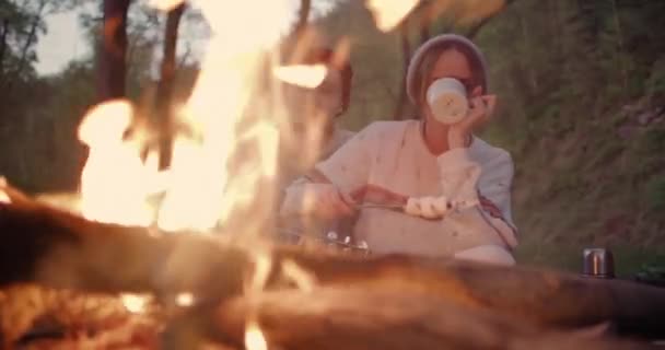 若いカップルが火のそばに座って話し、男がマシュマロをフライドポテトし、女の子がお茶を飲む. — ストック動画