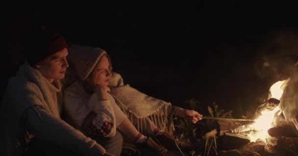 Una giovane coppia si siede di notte nella foresta, abbracciata sotto un plaid comune davanti a un falò, parlando e ridendo . — Video Stock