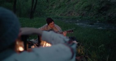 Genç bir adam bir dağ nehri yakınında bir ateşin önünde oynuyor, ve kız arkadaşı bir kamera ile onu fotoğraflıyor.