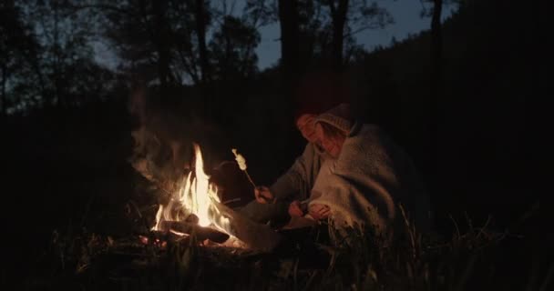 Una joven pareja se sienta por la noche en el bosque, abrazándose bajo un tartán común frente a una hoguera, hablando y riendo . — Vídeo de stock