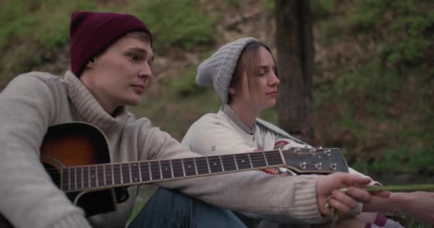 Kamarád s kytarou a přítelkyní sedí před ohněm v přírodě a baví se zábavou. — Stock video
