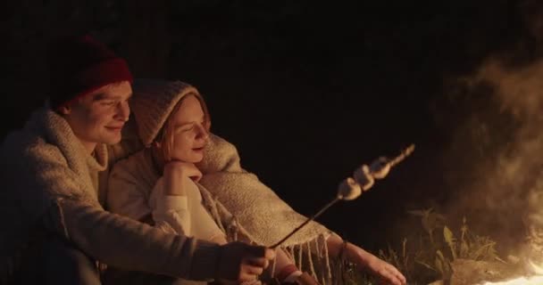 A young couple sits at night in the forest, embracing under a common plaid in front of a bonfire, talking and laughing. — Stock Video