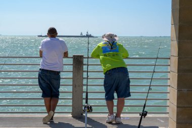 Fort De Soto Park, Florida -- February 17, 2019. Two fishermen on a pier pause to watch a large ship go by. clipart