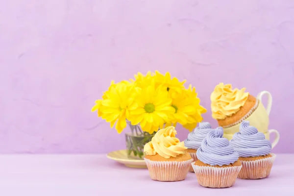 Cupcakes decorated with sweet yellow and violet cream and chrysanthemums on violet pastel background. Copyspace area. Can be used for greeting, mothers days and valentines card. Minimalism concept.