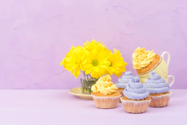 Cupcakes decorated with sweet yellow and violet cream and chrysanthemums on violet pastel background. Copyspace area. Can be used for greeting, mothers days and valentines card. Minimalism concept.