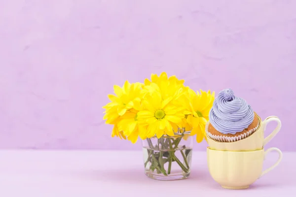 Cupcakes decorated with violet cream and bouquet of yellow chrysanthemums on violet pastel background. Text area. Ideal for greeting, mothers days and valentines card. Minimal still life concept.