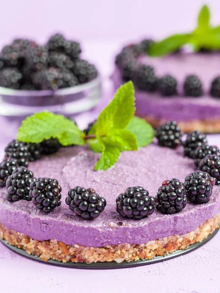 Raw blackberry dessert decorated with fresh ripe forest berries and green mint leaves on pastel violet background - sweet organic food for healthy eating and dieting concept.