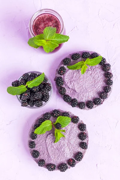 Raw blackberry dessert decorated with fresh ripe forest berries and green mint leaves and blended smoothie on pastel violet background - top view on sweet organic healthy food.