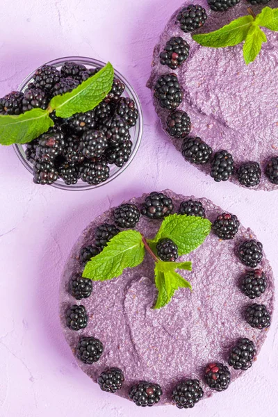 Raw blackberry dessert decorated with fresh ripe forest berries and green mint leaves on pastel violet background - sweet organic food for healthy eating and dieting concept.